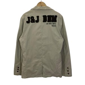 May include: Beige blazer with vertical stripes and long sleeves. The back of the jacket features the text "J&J DIM" in black, along with "SN-M8/NYC SS03". The blazer has three buttons on each cuff and a notched lapel.