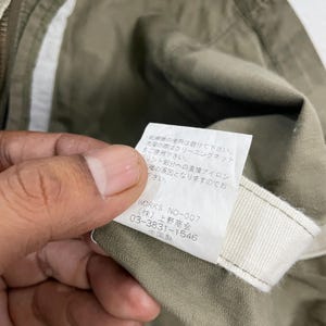 May include: Close-up of a tag with Japanese text and the text "WORKS NO-007" on a khaki-colored garment. The tag is white with black text and is held by a person's fingers.