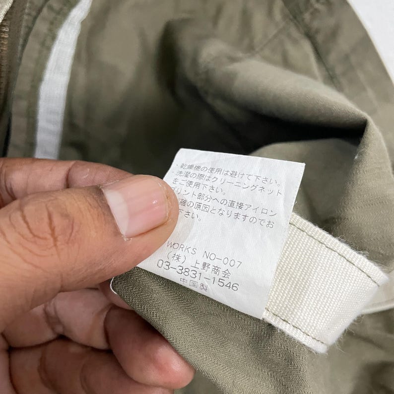 May include: Close-up of a tag with Japanese text and the text "WORKS NO-007" on a khaki-colored garment. The tag is white with black text and is held by a person's fingers.