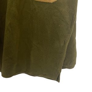 May include: Close-up of a dark olive green fleece fabric with a brown patch at the top. The fleece has a soft, textured appearance. The edges of the fabric are visible, with a small slit at the bottom.