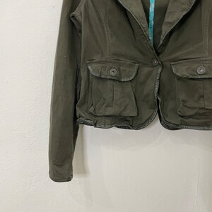 May include: A dark green cropped jacket with a collar, long sleeves, and two flap pockets with button closures. The jacket has a decorative trim around the bottom edge.