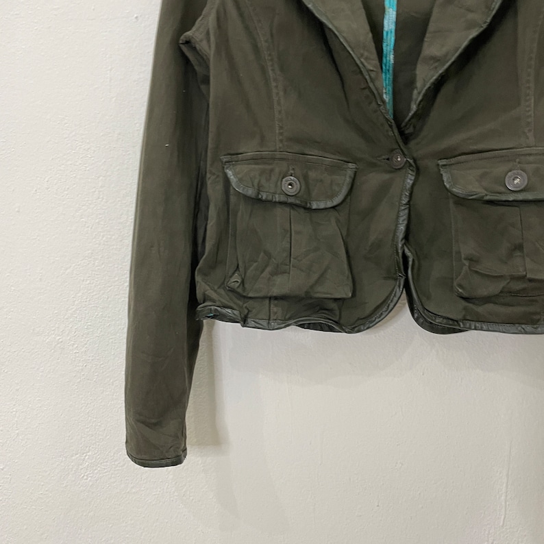 May include: A dark green cropped jacket with a collar, long sleeves, and two flap pockets with button closures. The jacket has a decorative trim around the bottom edge.