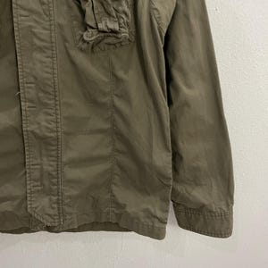 May include: Olive green jacket with a button-up front, a small pocket, and a collar. The jacket has a tailored fit and long sleeves. The fabric appears to be a lightweight material, suitable for layering. The jacket is a solid color.
