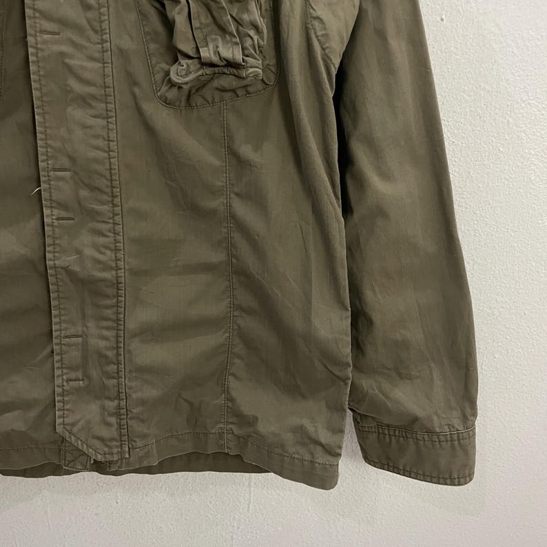 May include: Olive green jacket with a button-up front, a small pocket, and a collar. The jacket has a tailored fit and long sleeves. The fabric appears to be a lightweight material, suitable for layering. The jacket is a solid color.