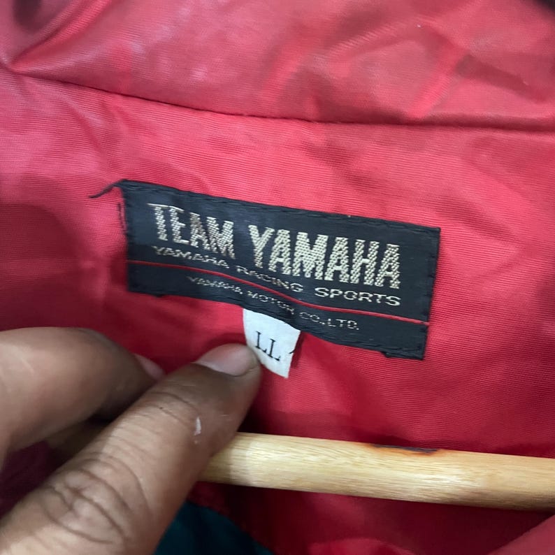K&ouml;nnte beinhalten: Nahaufnahme einer roten Jacke mit einem schwarzen Etikett mit der Aufschrift "TEAM YAMAHA" in wei&szlig;er Schrift. Das Etikett enth&auml;lt auch "YAMAHA RACING SPORTS" und "YAMAHA MOTOR CO., LTD." Ein wei&szlig;es Etikett mit "LL" ist angebracht.