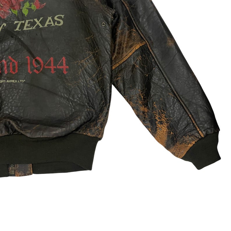 May include: A distressed dark brown leather bomber jacket with a black ribbed hem and cuffs. The front features a graphic of red roses, the text OF TEXAS in white, and the red text 1944, with the small white text COPYRIGHT AVIREX LTD visible.