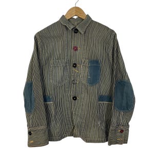 May include: A striped jacket with blue denim patches on the elbows and pocket. The jacket has a button-down front with black and red buttons. The cuffs also have red buttons. The jacket is hanging on a wooden hanger.