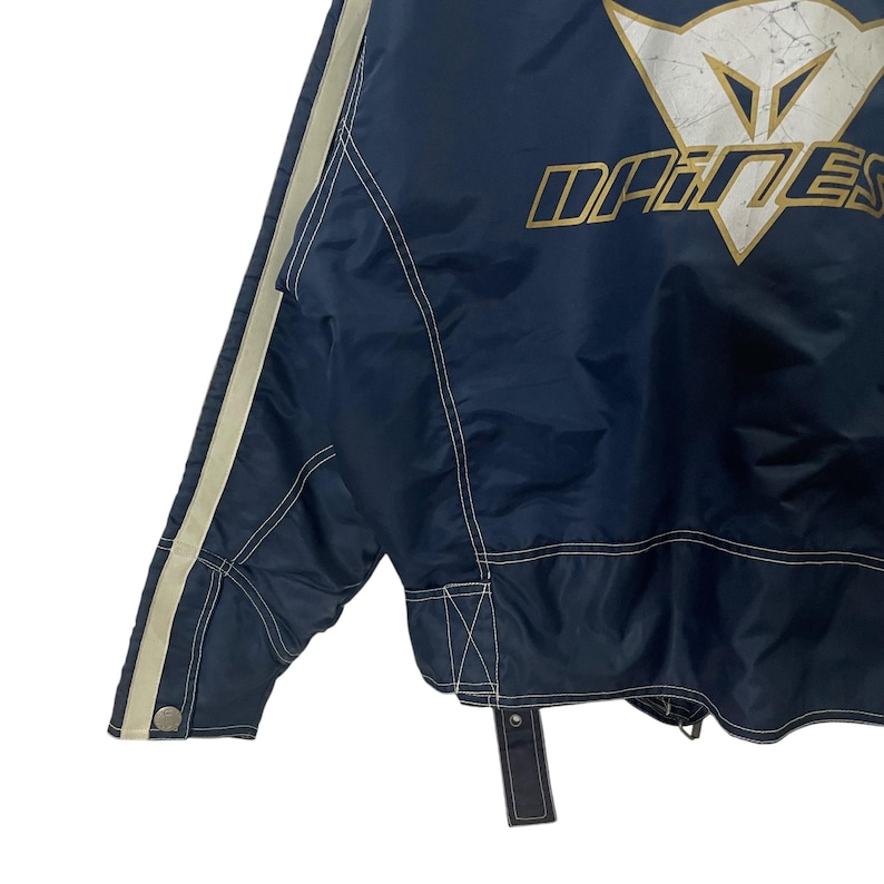 May include: A navy blue jacket with white piping and a DAINESE logo on the back. The jacket has a zipper closure and a white drawstring.