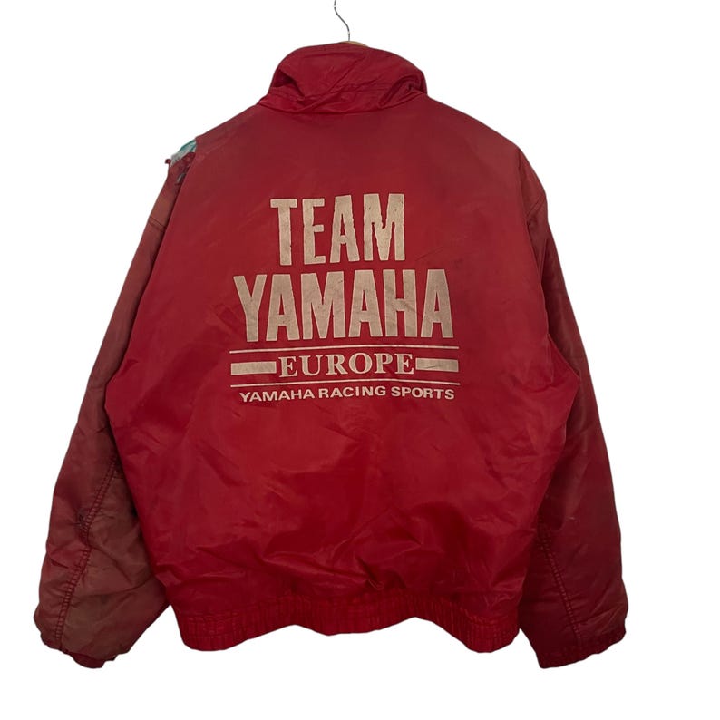 K&ouml;nnte beinhalten: Rote Team Yamaha Europe Rennjacke. Auf der Jacke steht in heller Farbe "TEAM YAMAHA EUROPE" auf dem R&uuml;cken. Die Jacke hat einen Kragen und einen Rei&szlig;verschluss.