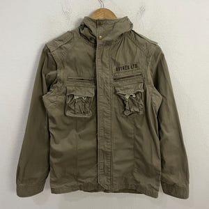 May include: Olive green jacket with multiple front pockets and a high collar. The jacket features the text "AVIREX LTD" on the upper chest. The jacket is hanging on a wooden hanger.
