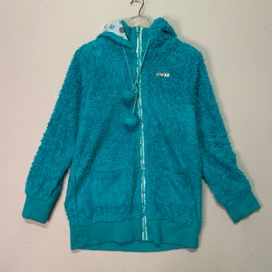 May include: A teal blue fleece hoodie with a white and green patterned hood and a zip closure. The hoodie has two pockets and a pom-pom detail on the zip pull.
