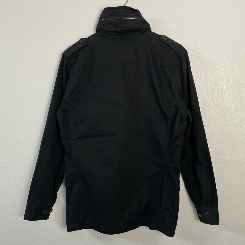 May include: A black jacket with a high collar and a zipper closure. The jacket has button details on the shoulders and cuffs. The back of the jacket is visible, showing the seams and construction.