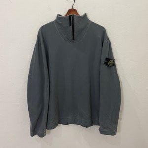 Puede incluir: Una sudadera de manga larga en color gris carbón con cuello alto y cierre de cremallera frontal en negro. La prenda incluye un parche con el logotipo en forma de brújula en negro y amarillo, sujeto a la manga izquierda con dos botones negros.
