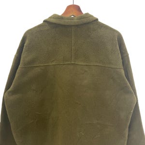May include: Olive green fleece jacket with a collared neckline and button closure. The back of the jacket is visible, showing the texture of the fleece material. The jacket is hanging on a wooden hanger.
