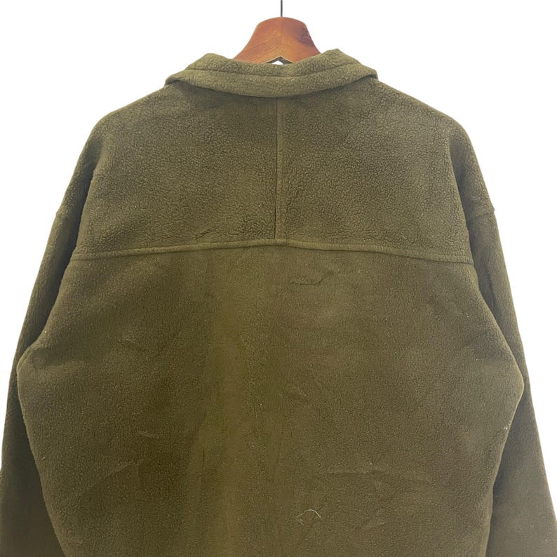 May include: Olive green fleece jacket with a collared neckline and button closure. The back of the jacket is visible, showing the texture of the fleece material. The jacket is hanging on a wooden hanger.