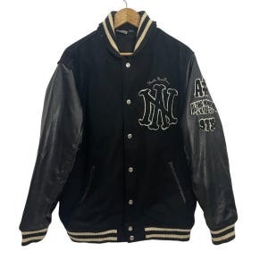 Könnte beinhalten: Schwarze Collegejacke mit Lederärmeln. Sie hat ein weißes "MN"-Emblem auf der Brust und "AE ALPHA NUMERIC 978" auf dem Ärmel. Kragen und Manschetten haben weiße und cremefarbene Streifen. Die Jacke ist zugeknöpft.