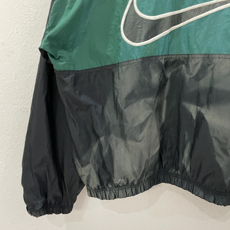 May include: A vintage Nike windbreaker jacket in green and black. The jacket features a large white-outlined Nike swoosh logo on the upper chest. The jacket has a drawstring hem and elastic cuffs.