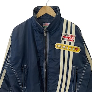 May include: A blue nylon jacket with white stripes and a Yamaha logo. The jacket has a zipper closure and a stand-up collar. The jacket also has a patch with the text "Motorcycle Top Fashion Leader NANKAI For Good Motorcycle Life".