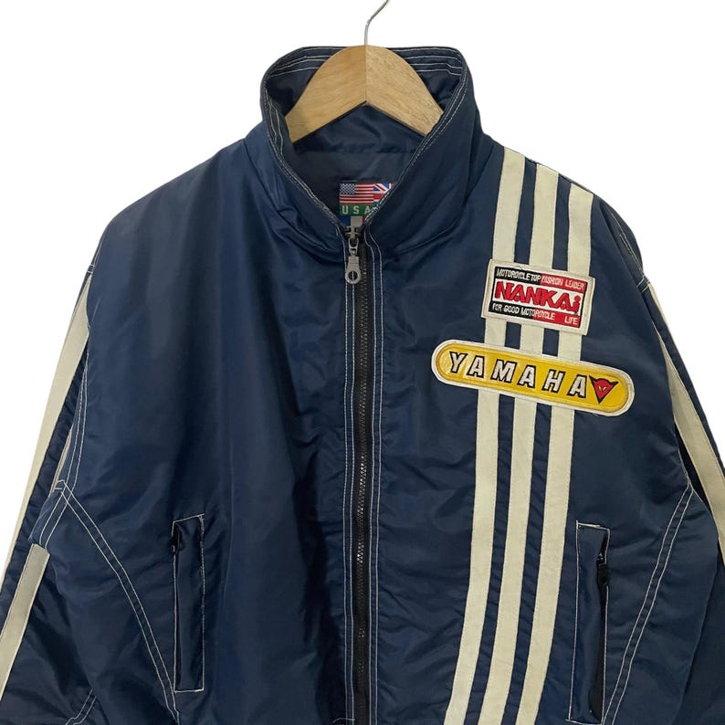 May include: A blue nylon jacket with white stripes and a Yamaha logo. The jacket has a zipper closure and a stand-up collar. The jacket also has a patch with the text "Motorcycle Top Fashion Leader NANKAI For Good Motorcycle Life".