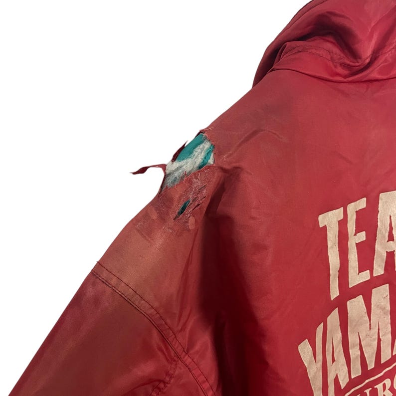 K&ouml;nnte beinhalten: Rote Vintage-Jacke mit wei&szlig;em Text "TEAM YAMAHA". Der &Auml;rmel der Jacke ist zerrissen und gibt einen t&uuml;rkisfarbenen und wei&szlig;en Innenraum frei. Die Jacke besteht aus einem leichten Material.