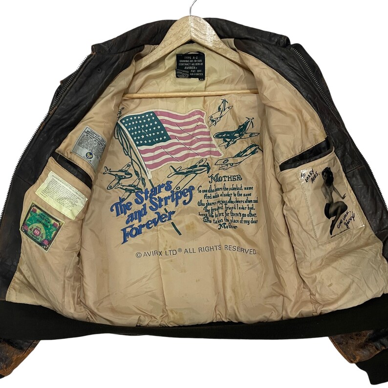 May include: A dark brown leather flight jacket with a beige interior lining. The lining features a printed American flag, illustrations of fighter planes, the text The Stars and Stripes Forever, a poem titled Mother, and a pin-up girl graphic.