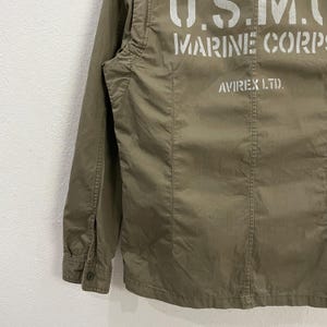 May include: Olive green military-style jacket with the text "U.S.M.C. MARINE CORPS" printed in white across the back. The jacket also features the text "AVIREX LTD.". The jacket has a buttoned cuff and a button closure.
