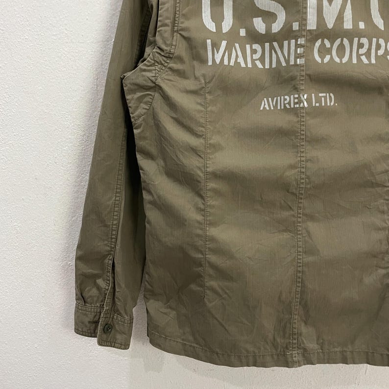 May include: Olive green military-style jacket with the text "U.S.M.C. MARINE CORPS" printed in white across the back. The jacket also features the text "AVIREX LTD.". The jacket has a buttoned cuff and a button closure.