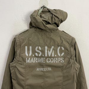 May include: Olive green jacket with a hood. The back of the jacket features the text "U.S.M.C. MARINE CORPS" in white block letters, with "AVIREX LTD." below. The jacket is hanging against a white wall.