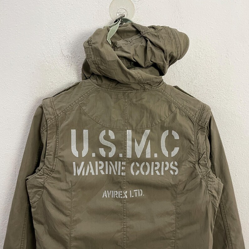 May include: Olive green jacket with a hood. The back of the jacket features the text "U.S.M.C. MARINE CORPS" in white block letters, with "AVIREX LTD." below. The jacket is hanging against a white wall.