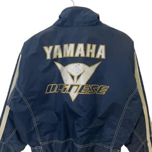 May include: A blue jacket with white and gold accents. The back of the jacket features a large white and gold Yamaha Dainese logo.