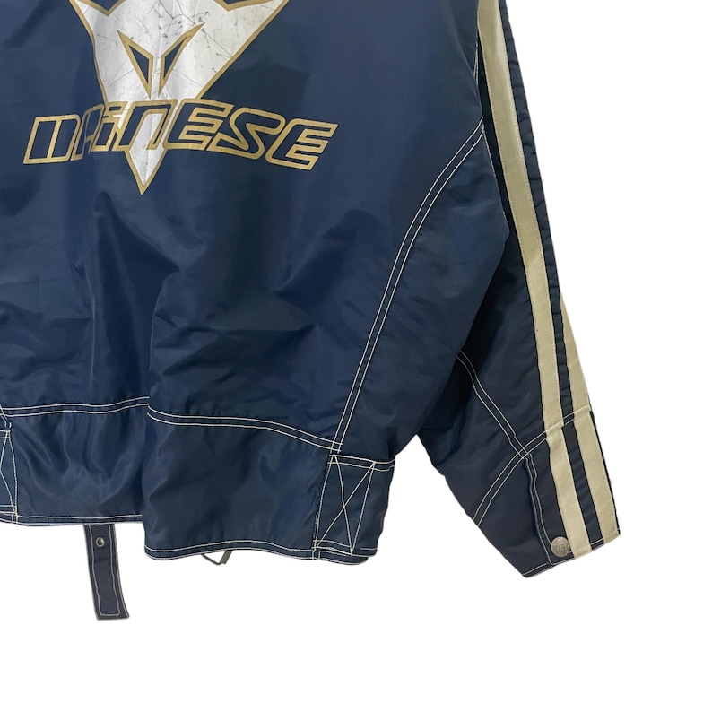 May include: A navy blue jacket with white stitching and white stripes on the sleeves. The jacket has a gold logo that says "Dainese" on the back.