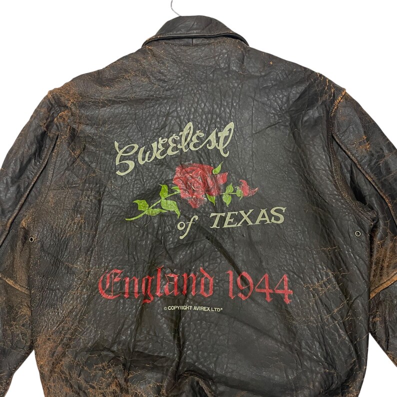 May include: A dark brown distressed leather jacket featuring a back graphic with a red rose and the text Sweetest of TEXAS and England 1944. The jacket has a collar, long sleeves, and the printed text COPYRIGHT AVIREX LTD near the bottom hem.
