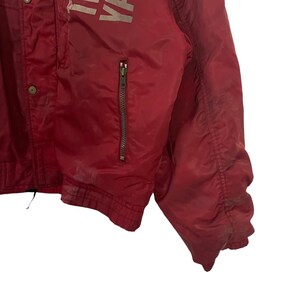 K&ouml;nnte beinhalten: Rote Vintage-Jacke mit Rei&szlig;verschlusstasche und Druckkn&ouml;pfen. Die Jacke hat verblasste wei&szlig;e Schrift auf dem oberen Teil. Die Jacke besteht aus einem gl&auml;nzenden Material und weist Gebrauchsspuren auf.