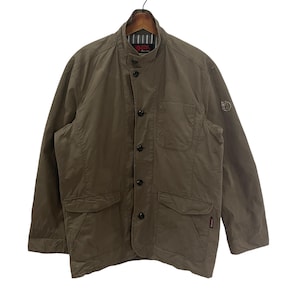 May include: A brown utility jacket with a button-up front, two large pockets, and a smaller pocket on the chest. The jacket has a stand-up collar and a logo on the upper right sleeve.
