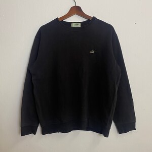 Könnte beinhalten: Schwarzer Langarm-Pullover mit einem kleinen gestickten Logo auf der linken Brust. Das Logo ist ein stilisiertes Bild eines Krokodils.