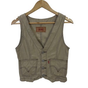 Può includere: Gilet in denim beige con scollo a V, due tasche frontali e chiusura con bottoni. Presenta cuciture scure e una piccola etichetta rossa. Il gilet è appeso a una gruccia di legno.