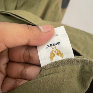 Op de afbeelding: Close-up van een olijfgroen kledingstuk met een wit label. Het label toont de merknaam "AVIREX" in zwart, met een gouden embleem van een ster en vleugels. De stof van het kledingstuk heeft een subtiele textuur.