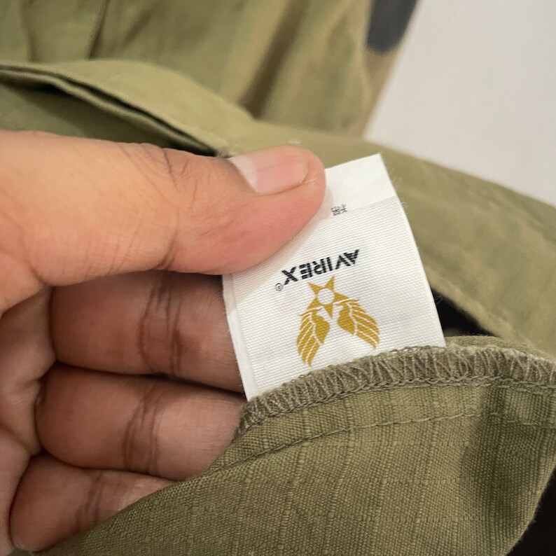 Op de afbeelding: Close-up van een olijfgroen kledingstuk met een wit label. Het label toont de merknaam "AVIREX" in zwart, met een gouden embleem van een ster en vleugels. De stof van het kledingstuk heeft een subtiele textuur.