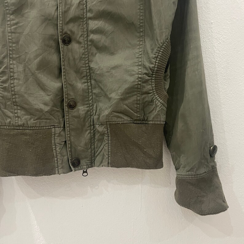 May include: A green utility jacket with a zipper closure and button accents. The jacket has a ribbed waistband and cuffs.