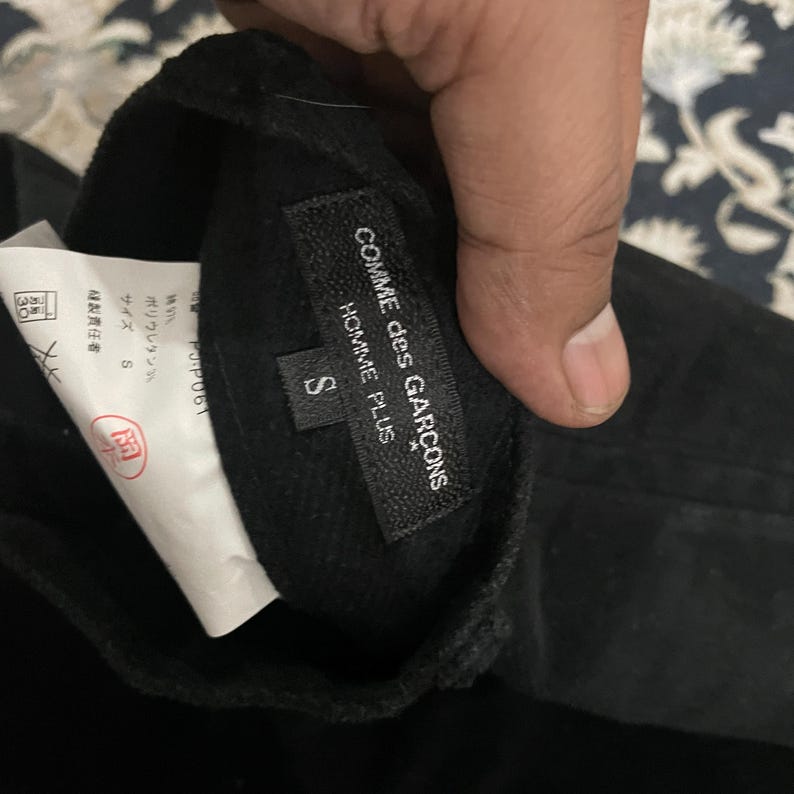 Puede incluir: Etiqueta negra Comme des Gar&ccedil;ons Homme Plus con texto blanco. La etiqueta dice "COMME des GAR&Ccedil;ONS HOMME PLUS" y "S".
