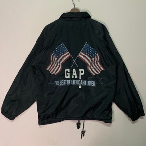 Puede incluir: Chaqueta cortavientos negra con cierre de cremallera y cordón de ajuste en la parte inferior. La parte posterior de la chaqueta presenta un gráfico de dos banderas americanas cruzadas con la palabra "GAP" y el texto "The Best of American Flavor".
