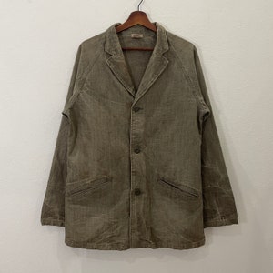 May include: A long-sleeve olive green jacket with a notched lapel collar, three front buttons, and two slanted welt pockets at the waist. The garment features raglan sleeves and a textured fabric finish, displayed on a wooden hanger.