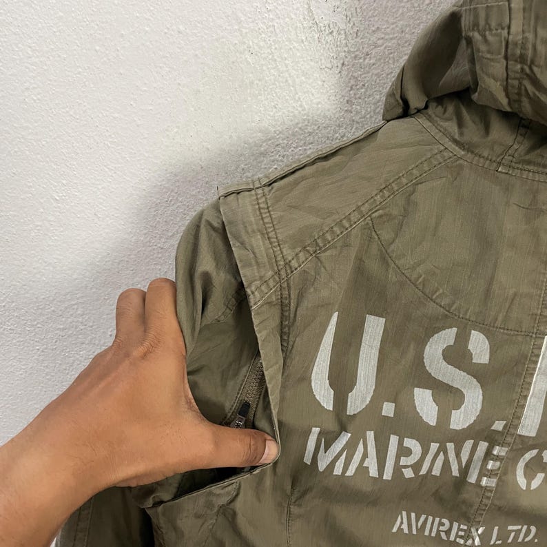 May include: Olive green jacket with a hood and the text "U.S. MARINE CORPS" printed in white. The jacket has a zipper pocket and the brand name "AVIREX LTD." is visible.