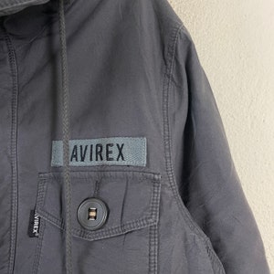 Pode incluir: Close-up de um casaco cinza carv&atilde;o com capuz e bolso frontal. O casaco apresenta um patch retangular com a palavra "AVIREX" em letras cinzentas. Uma fecho de bot&atilde;o &eacute; vis&iacute;vel no bolso.