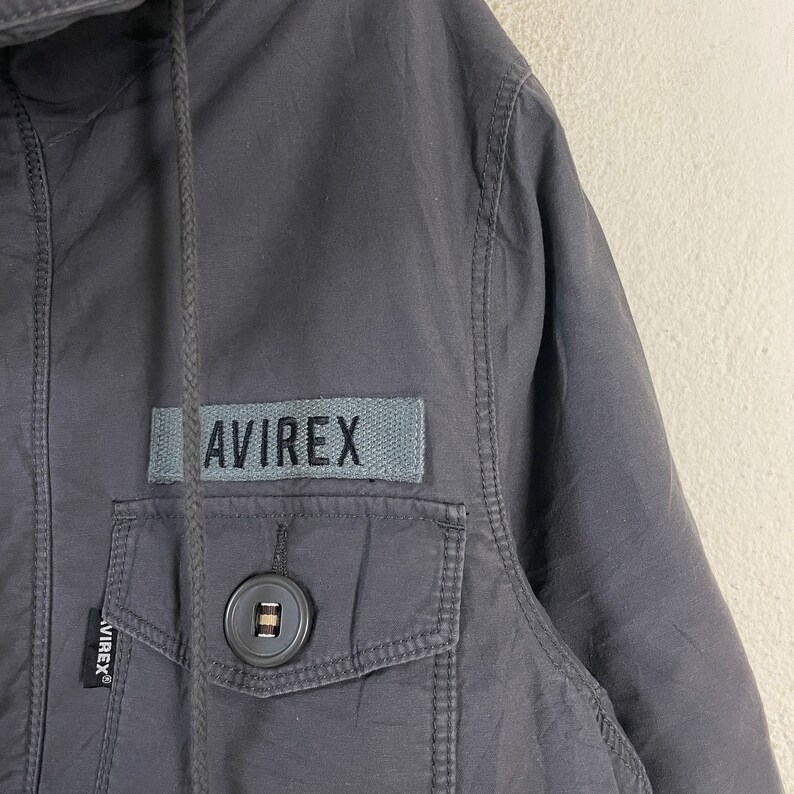 Pode incluir: Close-up de um casaco cinza carv&atilde;o com capuz e bolso frontal. O casaco apresenta um patch retangular com a palavra "AVIREX" em letras cinzentas. Uma fecho de bot&atilde;o &eacute; vis&iacute;vel no bolso.