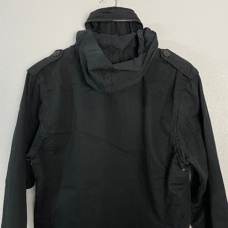 May include: Black hooded jacket with a zipper closure and button details on the shoulders. The jacket has a hood and a simple, utilitarian design. The jacket is made of a durable fabric.