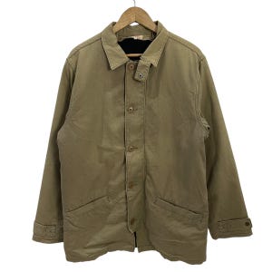 Puede incluir: Una chaqueta de color beige con cierre de botones, dos bolsillos delanteros y un cuello. La chaqueta está colgada de una percha de madera. La chaqueta tiene un forro oscuro y puños abotonados. La chaqueta parece estar hecha de una tela duradera.