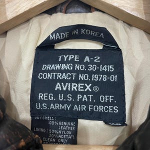 May include: A black rectangular garment label with white text reading TYPE A-2, DRAWING NO. 30-1415, CONTRACT NO. 1978-01, AVIREX, REG. U.S. PAT. OFF., U.S. ARMY AIR FORCES, MADE IN KOREA, 100% GENUINE LEATHER, 50% NYLON, 50% ACETATE.