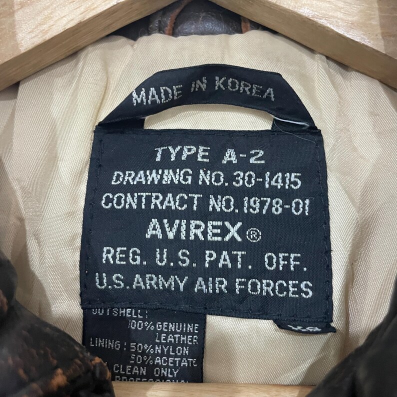 May include: A black rectangular garment label with white text reading TYPE A-2, DRAWING NO. 30-1415, CONTRACT NO. 1978-01, AVIREX, REG. U.S. PAT. OFF., U.S. ARMY AIR FORCES, MADE IN KOREA, 100% GENUINE LEATHER, 50% NYLON, 50% ACETATE.