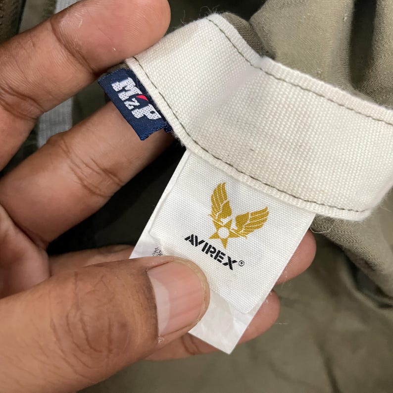 May include: Close-up of a garment label with the brand name AVIREX and a gold emblem of a stylized eagle. The label is white with black text and a gold emblem. The garment is olive green.